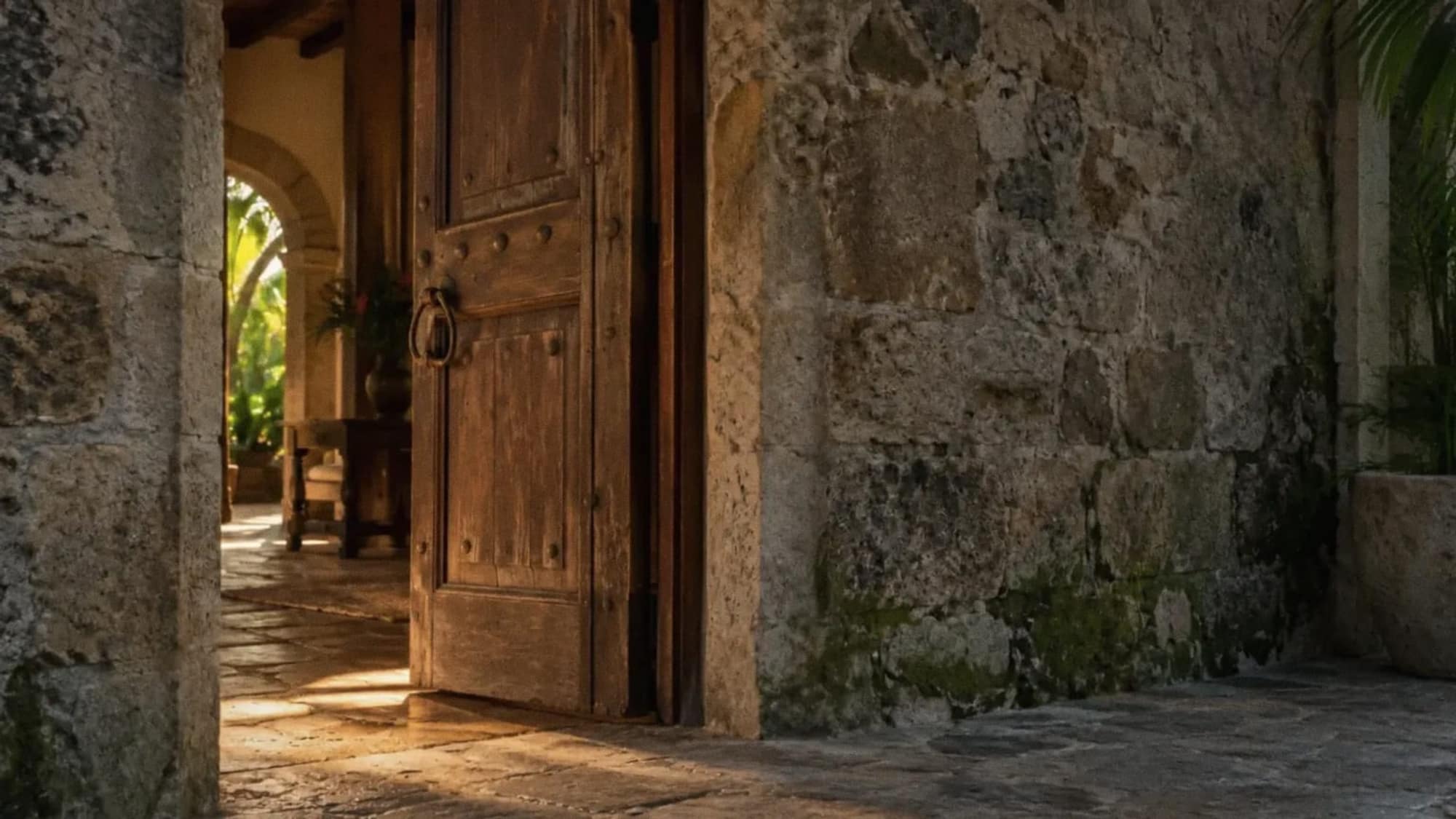 Puerta antigua de madera entreabierta en un muro de piedra; adentro, un corredor con luz de tarde y un patio con plantas.